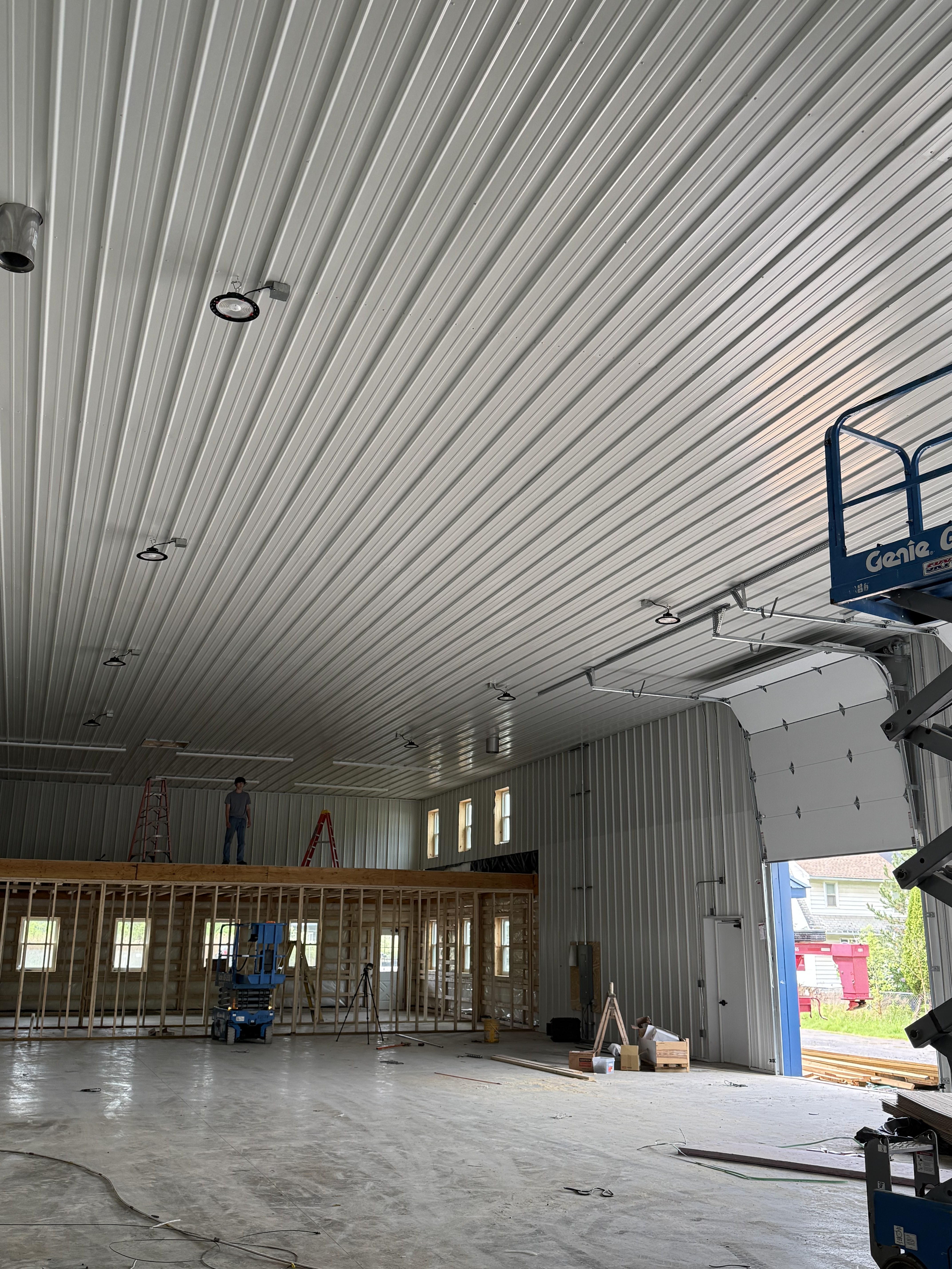 Commercial metal building under construction with electrician on ladder installing high-bay LED lights, Genie scissor lift โ Monroe County NY
