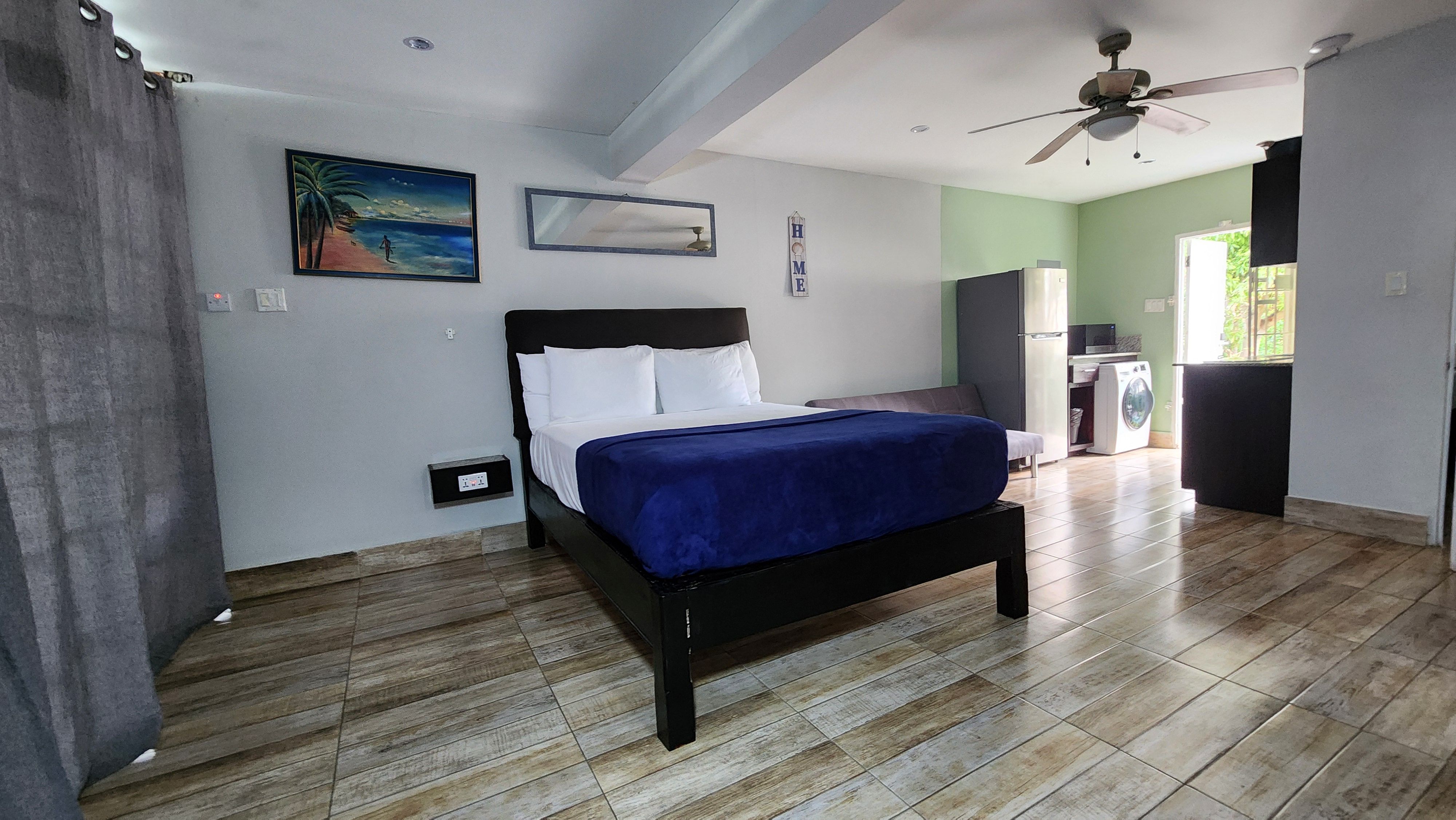 MG17 studio — queen bed, Jamaican beach artwork, HOME sign, kitchen with fridge and washer visible