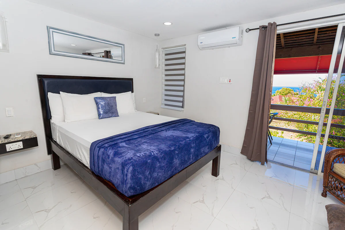 Bedroom — queen bed with dark platform frame, navy blue bedding, blue sequin pillow, pendant lamp, zebra blinds, AC unit, sliding glass door with balcony and Caribbean Sea visible, marble tile floors