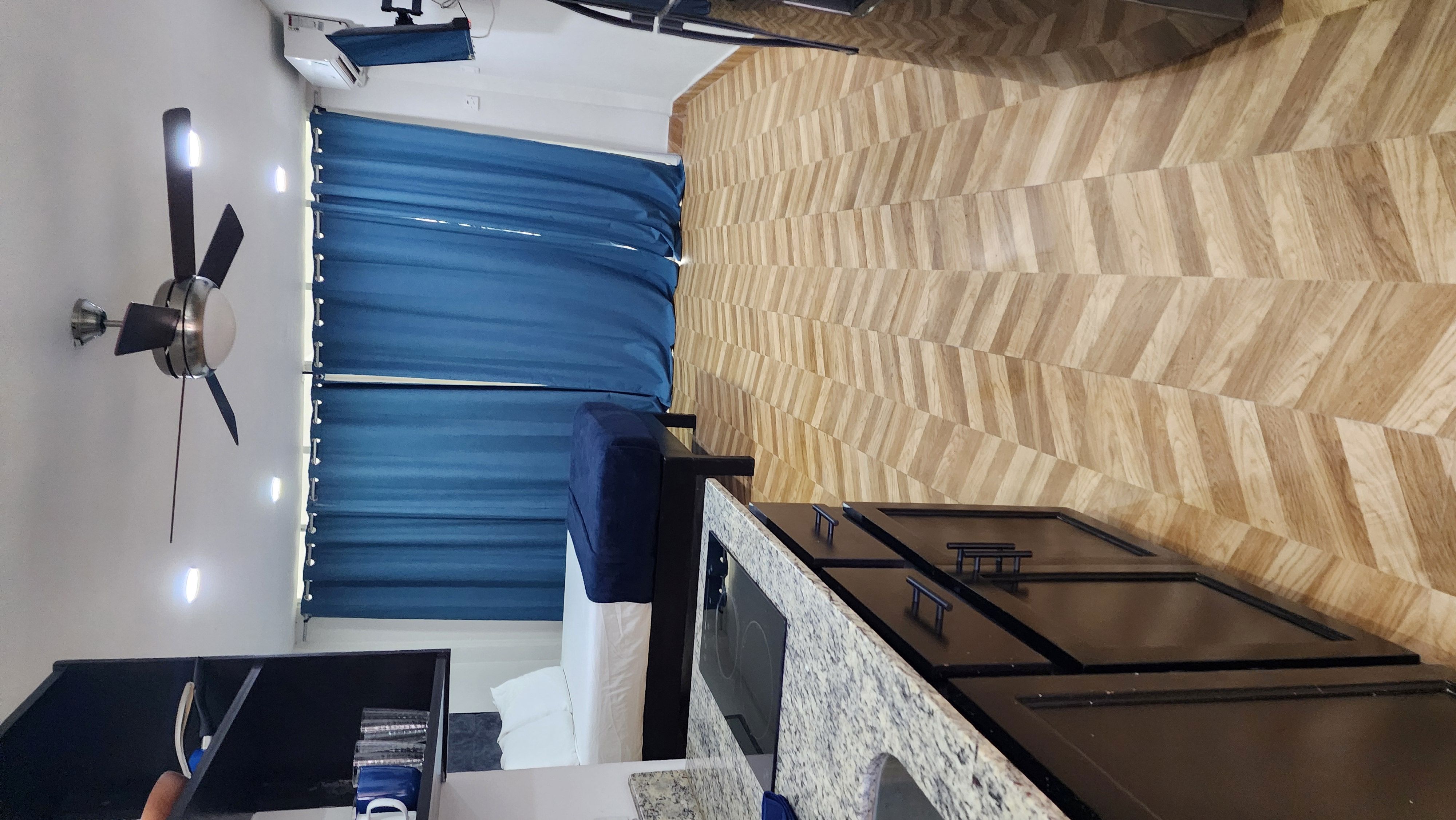 MG6 studio — queen bed, AC, ceiling fan, blue curtains, herringbone floors, granite kitchen counter