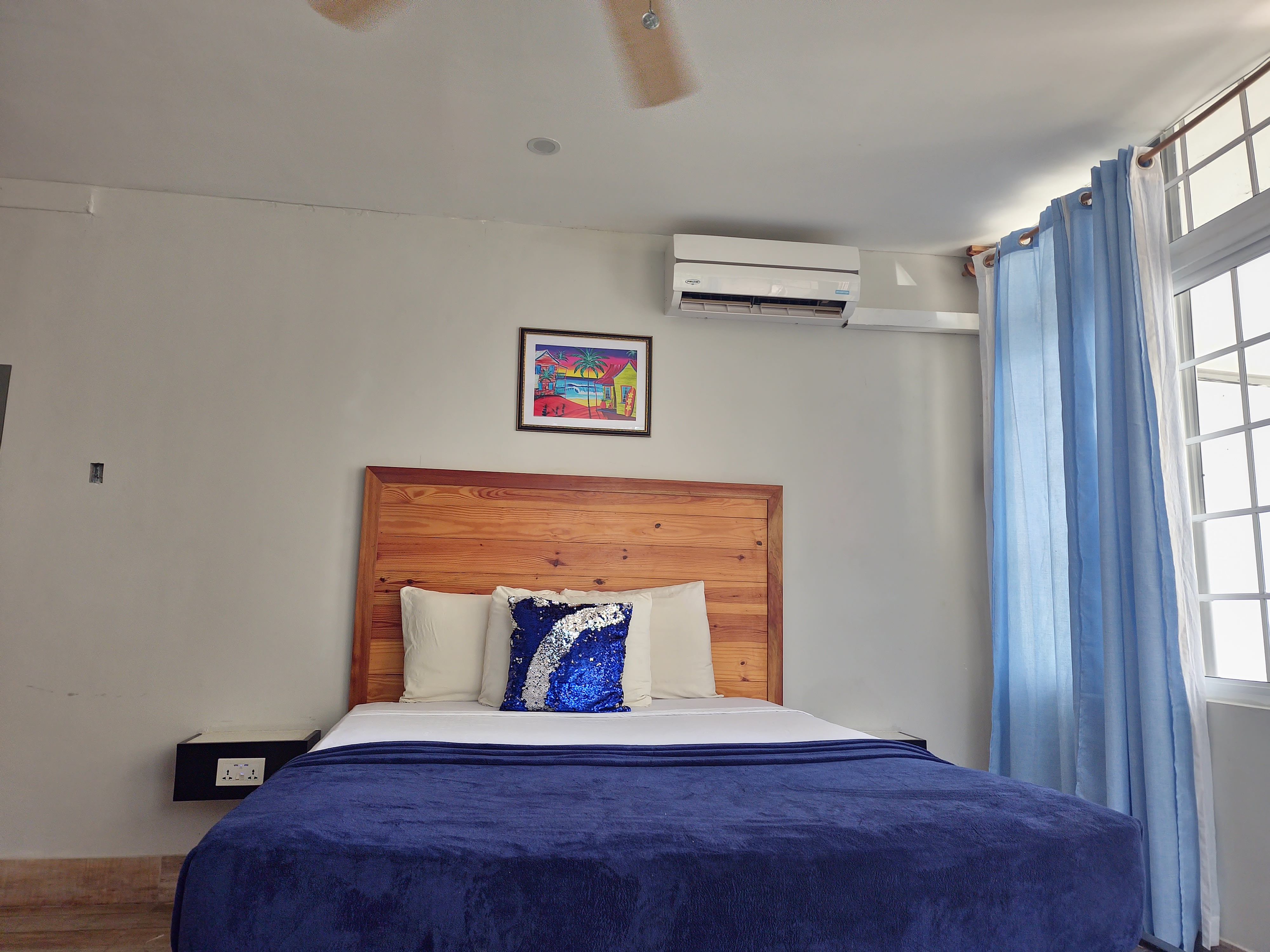 MG1 queen bed with AC unit, blue curtains, and Jamaican artwork