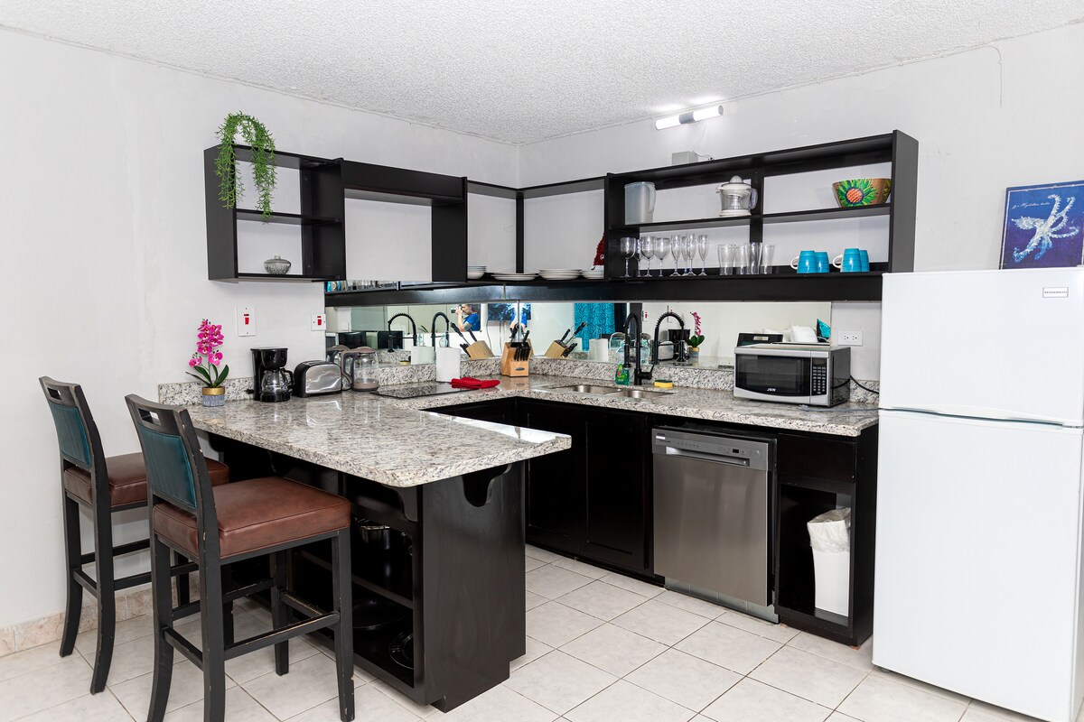 Full kitchen — dark cabinets, granite counters, breakfast bar with bar stools, open shelves with glassware, coffee maker, toaster, microwave, dishwasher, white fridge, mirrored backsplash