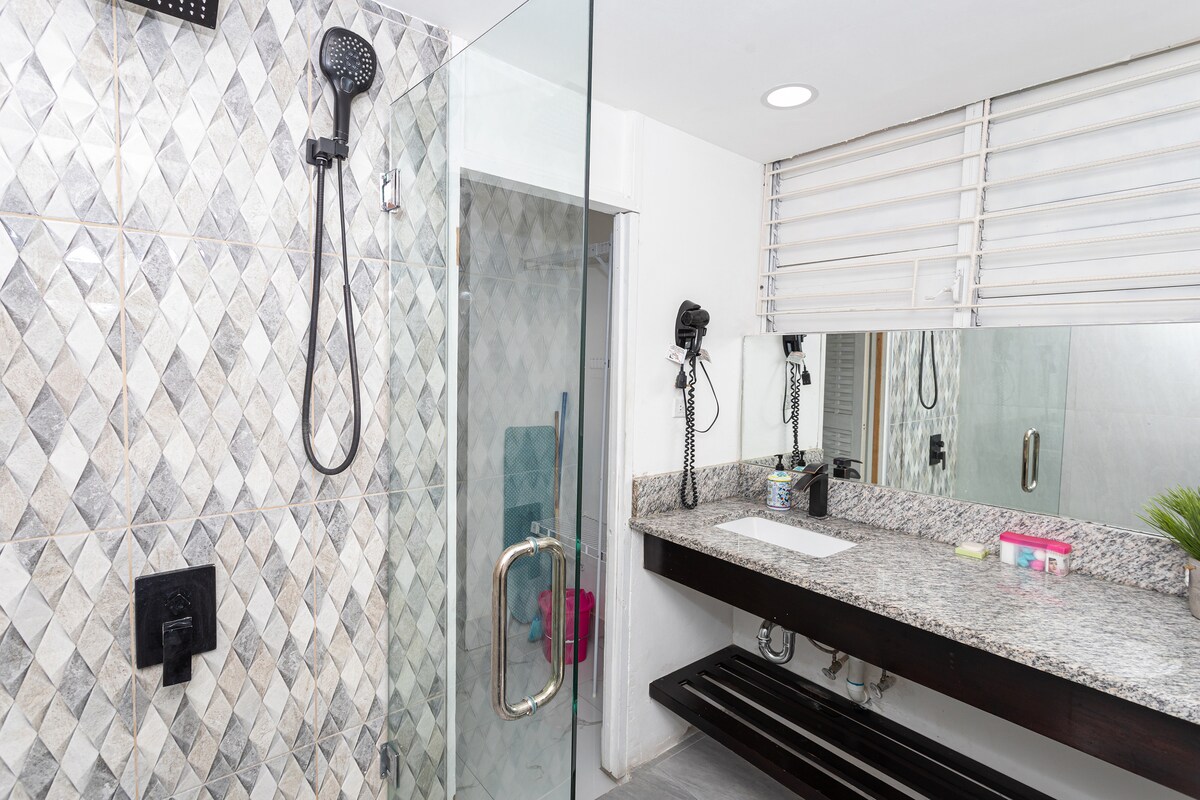 Bathroom — glass walk-in shower with diamond-pattern tiles, black fixtures and handheld showerhead, granite vanity, large mirror, hair dryer, white louvered window
