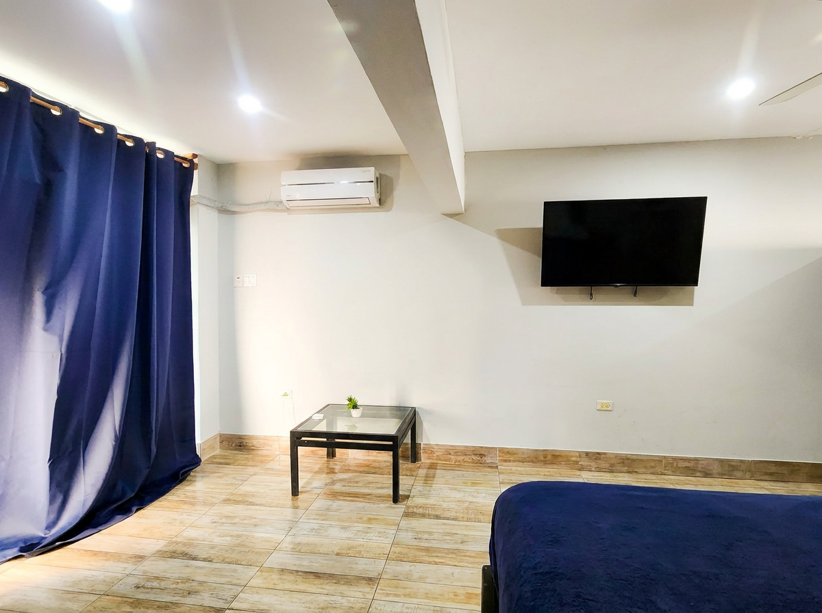 MG10 living area — wall-mounted Smart TV, AC unit, glass coffee table, navy blue curtains, tiled floor