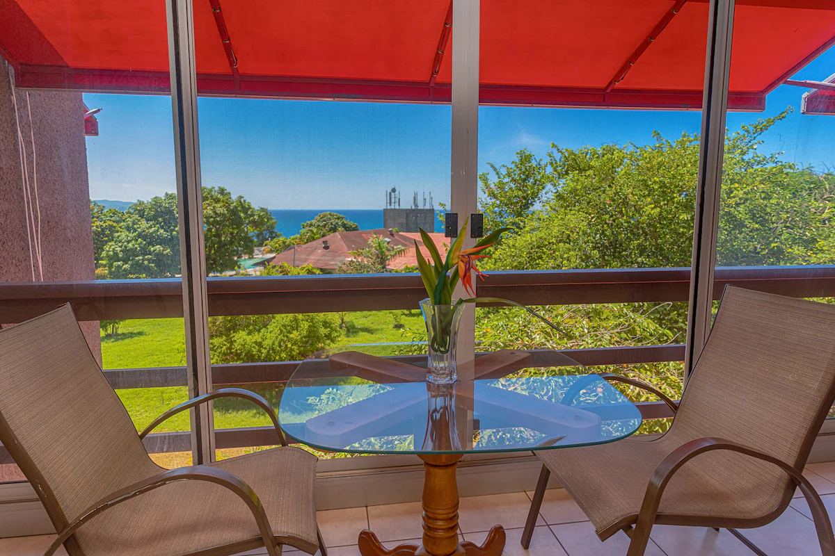 Covered balcony — round glass table with chairs under red awning, lush tropical garden and Caribbean Sea visible in the distance, birds of paradise flower