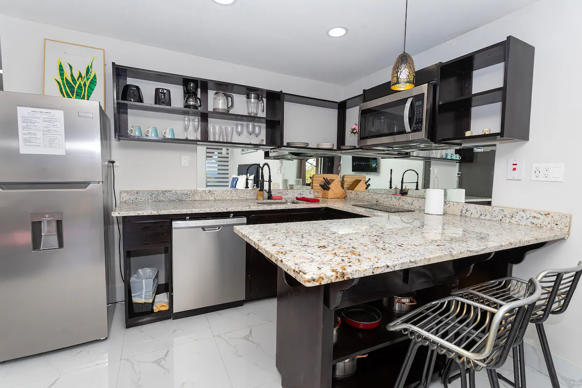 Full kitchen — dark cabinets, granite counters and breakfast bar, stainless fridge and dishwasher, microwave, open shelves with glassware and coffee maker, pendant light, mirrored backsplash, marble tile floors