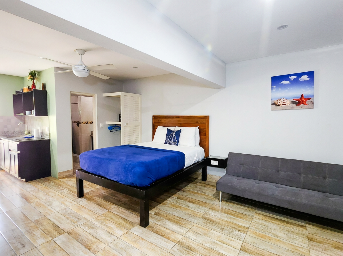 MG10 studio interior — queen bed with blue bedding, futon sofa, full kitchen, ceiling fan, beach artwork