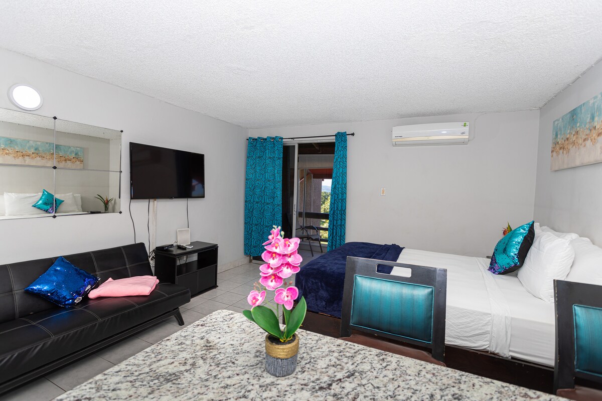 Studio wide angle — king bed with dark headboard, navy blue bedding, teal sequin pillows, black leather futon, wall-mounted TV, AC unit, teal curtains, beach artwork, granite breakfast bar