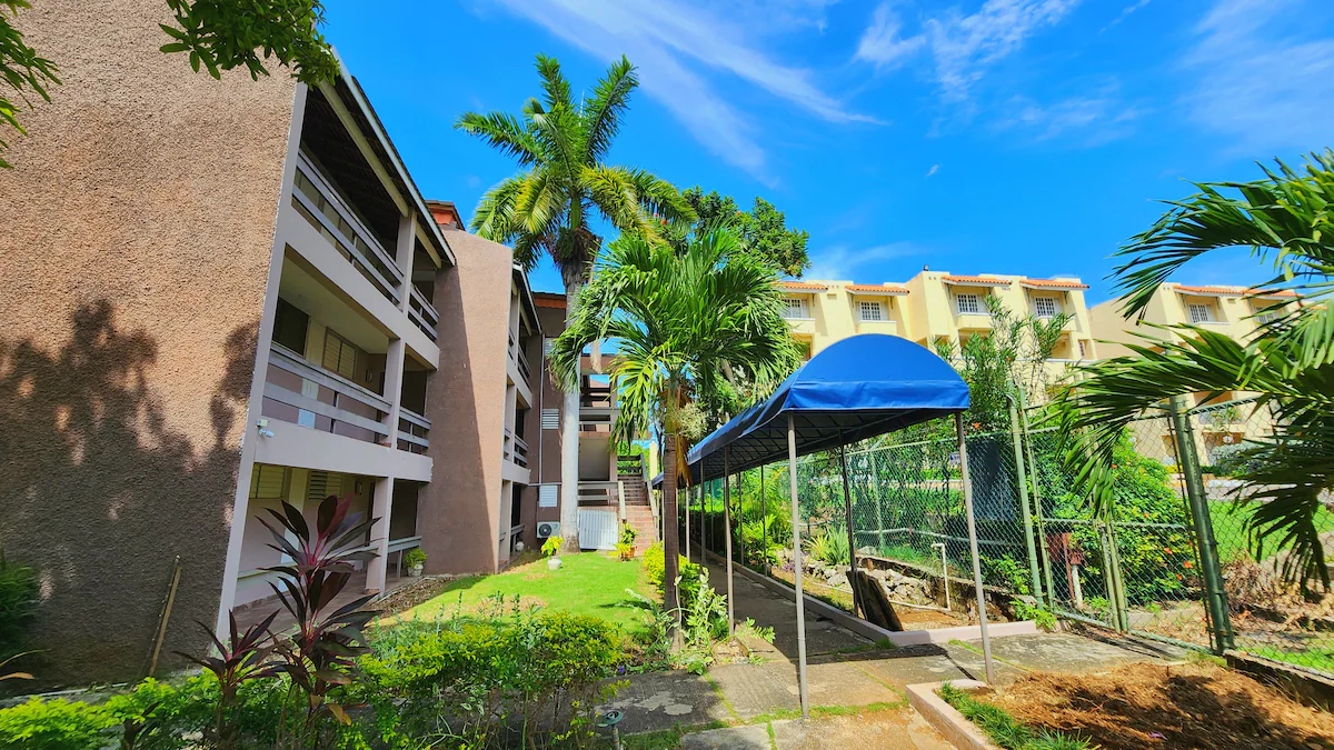 Garden Studios building exterior — 3-storey building with lush tropical garden, palm trees, blue sky, covered parking area
