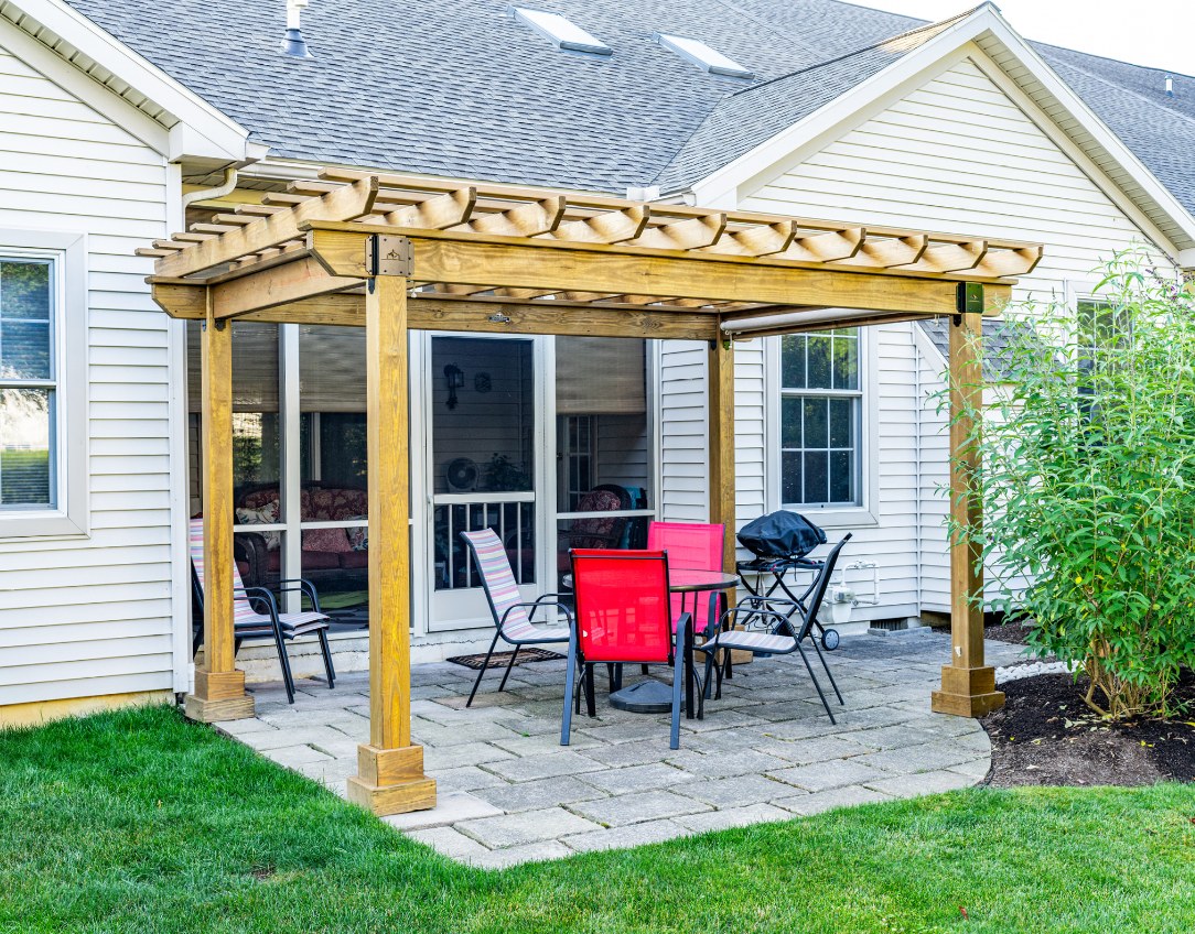 Golden-stained wood Hollander pergola on a paver patio beside a house, with red chairs and a dining table underneath