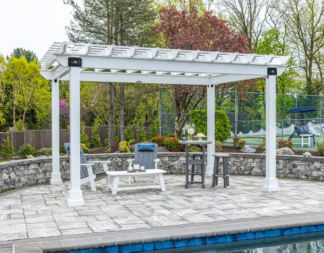 White vinyl Hollander pergola with black brackets on a stone paver patio beside a pool, with gray and white chairs and a bar-height table underneath