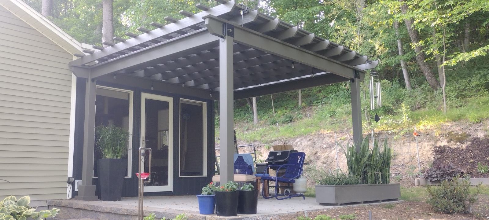 Gray-stained wood Hollander pergola with string lights in front of a black-sided room