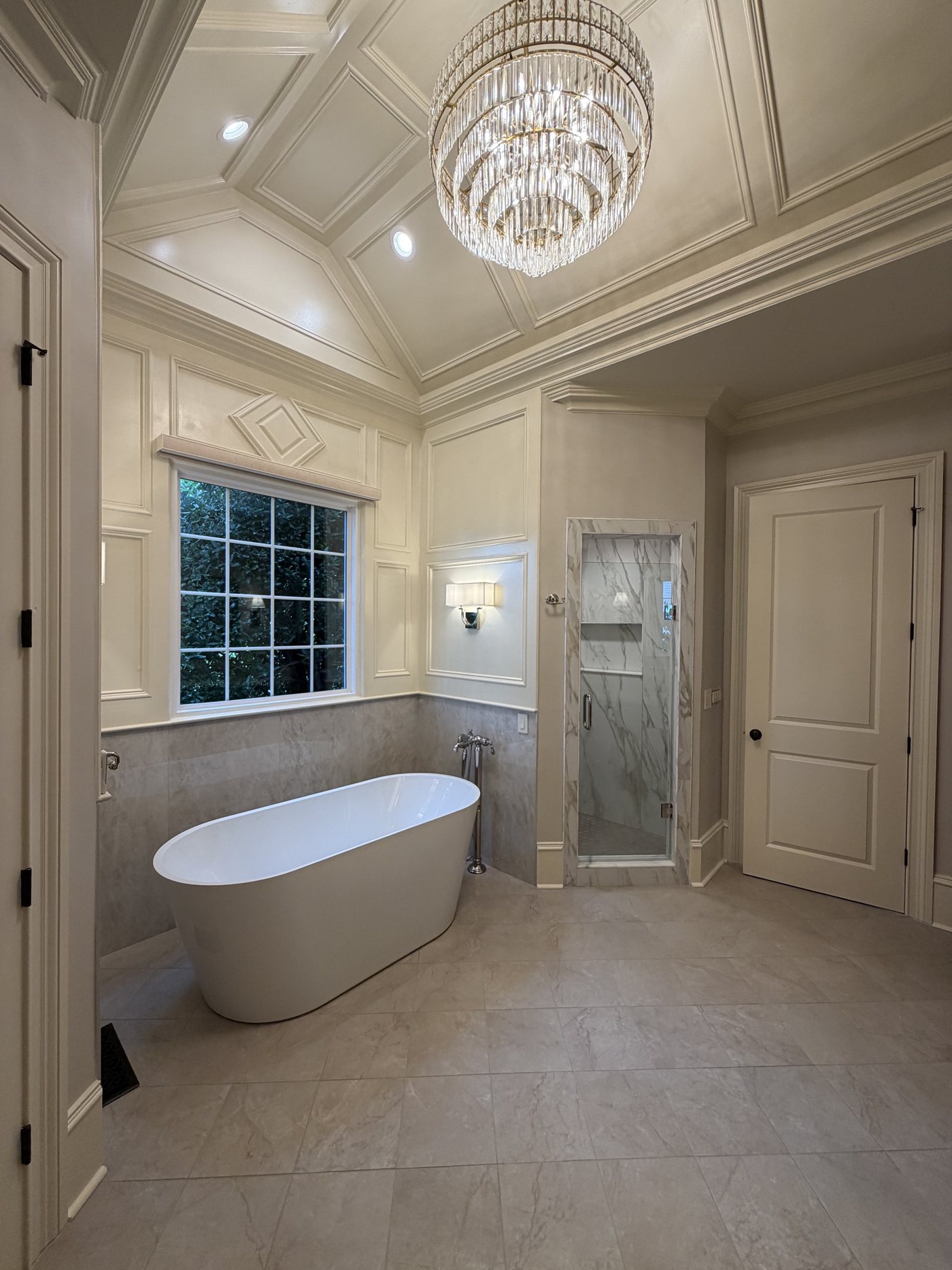 Freestanding soaking tub with crystal chandelier, paneled walls and marble-look shower